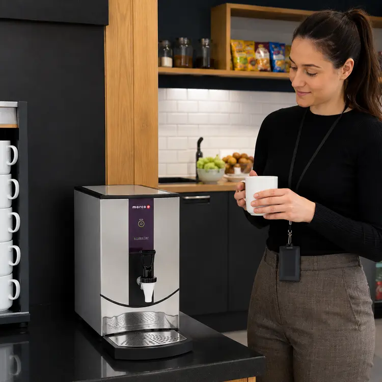 Person holding a white mug under a Marco EcoBoiler T5 hot water boiler in a modern office kitchen with shelves of mugs, snacks, and a tiled backsplash.