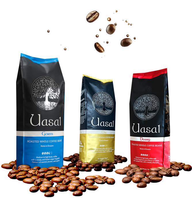 A dramatic studio shot of three 1kg bags of Uasal Roasted Whole Coffee Beans (Gorm, Óir, and Dearg) on a black background, with whole coffee beans levitating in the air above them.