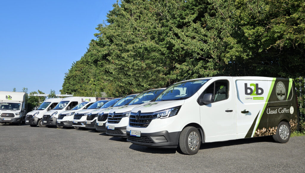 A row of professional white service vans parked outdoors, featuring the green "b2b Coffee Solutions" and "Uasal Coffee" branding on their sides.